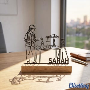 Op de afbeelding: Een zwart metalen architectonisch ontwerp met een figuur met een helm, die een blauwdruk meet. Een kraan, landmeetkundige apparatuur en de naam "SARAH" zijn ook inbegrepen. Het ontwerp is gemonteerd op een houten voet.