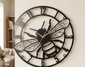 May include: Black metal wall clock featuring a bee design. The clock has Roman numeral hour markers and gold-colored clock hands. The bee's wings and body are intricately detailed.