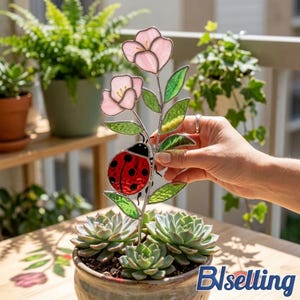 May include: A stained glass garden stake featuring a ladybug and pink flowers. The ladybug has a red body with black spots. The flowers and leaves are green and pink. The stake is held in a pot of succulents.