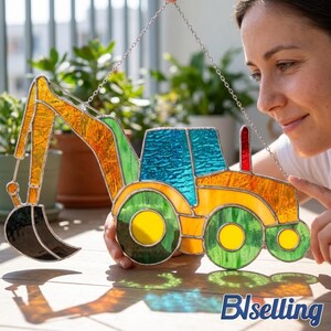 Op de afbeelding: Een kleurrijk glas-in-lood raamwerk van een graafmachine met gele, oranje, groene, blauwe en rode accenten. De graafmachine wordt opgehangen door een zilveren ketting. Het ontwerp omvat een bak, cabine en wielen.