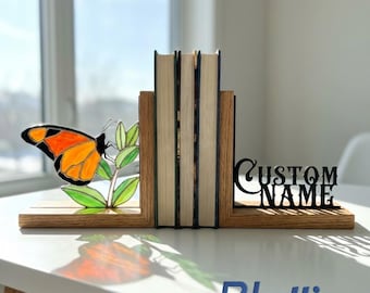 May include: Wooden bookends with a butterfly design and the text "CUSTOM NAME". The butterfly is orange and black with green leaves. The bookends are holding books on a white surface.