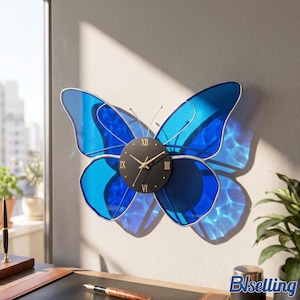 May include: A butterfly-shaped wall clock featuring translucent blue wings with silver-toned outlines. The central circular clock face is black with gold-colored Roman numerals and thin gold hands. Two thin metal antennae extend from the top.