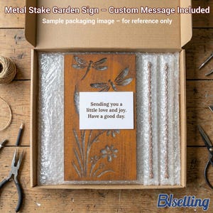 a wooden garden sign with a message, placed in a cardboard box with bubble wrap, and various crafting tools and materials are scattered around the box on a wooden surface.