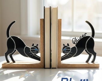 Puede incluir: Un par de sujetalibros de madera con siluetas de gatos negros, ojos azules y contornos plateados. El gato de la derecha tiene el nombre Oliver escrito en letra cursiva negra. Los sujetalibros están colocados a ambos lados de dos libros.