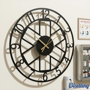 May include: A large, round, black metal wall clock with gold-colored hands. The clock face features large, stylized numerals and a geometric design. The clock is mounted on a white wall.