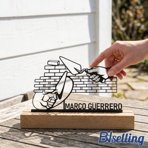 Puede incluir: Escultura de albañil de metal negro con el nombre "MARCO GUERRERO" sobre una base de madera. La escultura representa manos usando una llana para construir un muro de ladrillos. Es un artículo decorativo.