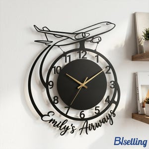 May include: Black metal wall clock featuring an airplane design above a circular clock face. The clock has gold-colored hands and the text "Emily's Airways" is written below the clock face. The clock is mounted on a white wall.