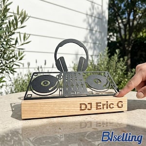 Puede incluir: Escultura de equipo de DJ de metal negro con auriculares, tocadiscos y mezclador, montada sobre una base de madera clara. El texto "DJ Eric G" está grabado en la base. La escultura se exhibe al aire libre.