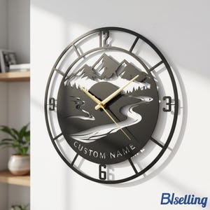 Puede incluir: Reloj de pared de metal negro con un paisaje de montaña y río. La esfera del reloj presenta un diseño recortado con una silueta de paisaje, manecillas doradas y el texto "CUSTOM NAME". El reloj está montado en una pared blanca.