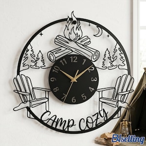 May include: Black metal wall clock with a camping theme. The clock features a circular design with a campfire, two Adirondack chairs, trees, and the words "Camp Cozy." The clock face is black with gold hands and numbers.