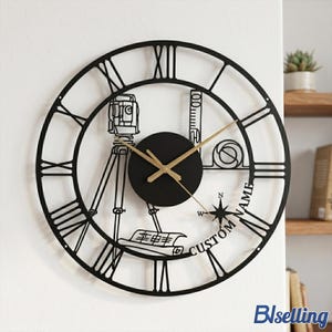 Peut inclure: Horloge murale en métal noir avec des chiffres romains et des aiguilles dorées. L'horloge présente un design circulaire avec des outils d'architecture et le texte "CUSTOM NAME". L'horloge est fixée sur un mur blanc.