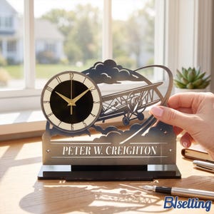 May include: A dark metal desk clock featuring a cutout design of a biplane flying through clouds. The clock face is circular with a black center, cream-colored border, and gold hands. The name PETER W. CREIGHTON is engraved on a horizontal bar below.