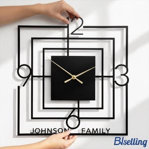 Op de afbeelding: Zwarte vierkante wandklok met gouden wijzers en de tekst "JOHNSON FAMILY". De klok heeft een uniek geometrisch ontwerp met de cijfers 2, 3, 6 en 9. De klok wordt door twee handen vastgehouden.