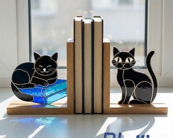 Puede incluir: Un par de sujetalibros de madera con gatos de vidrieras negras. Un gato está acostado sobre un libro de cristal azul, mientras que el otro está sentado. Los sujetalibros sostienen tres libros, con una ventana visible al fondo.
