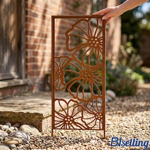 A hand is holding a decorative metal screen with a floral pattern, placed on a gravel path in a garden setting.