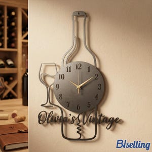 Op de afbeelding: Een grijze metalen wandklok in de vorm van een wijnfles en glas. De klok heeft een ronde wijzerplaat met gouden wijzers en zwarte cijfers. De woorden "Olivia's Vintage" staan eronder.