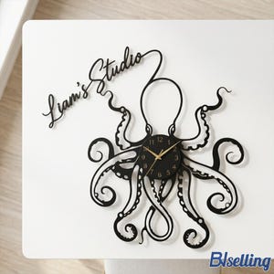 Op de afbeelding: Zwarte octopusvormige wandklok met gouden wijzers en cijfers. De klok heeft de tekst "Liam's Studio" in een cursief lettertype. De klok is gemonteerd op een wit oppervlak.