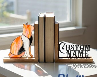 Puede incluir: Un par de sujetalibros de madera con una figura de gato de vidrieras en naranja, blanco y negro en un lado y un recorte de metal negro con el texto CUSTOM NAME en el otro, sosteniendo dos libros en posición vertical sobre una superficie blanca.