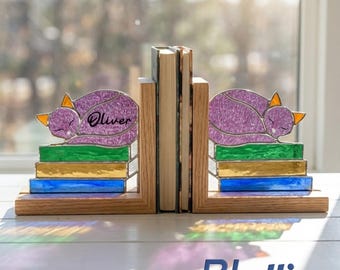 Puede incluir: Un par de sujetalibros de madera con figuras de gatos de vidrieras descansando sobre libros apilados. Los gatos son morados con orejas naranjas, y los libros son verdes, amarillos y azules. El gato izquierdo tiene el nombre Oliver.
