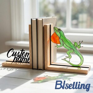 May include: Wooden bookends with a green and orange stained glass lizard design. The bookends hold several books and feature the text "Custom name". The lizard has a vibrant orange throat and is positioned as if climbing.