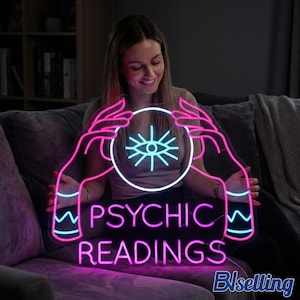May include: A neon sign with the words "PSYCHIC READINGS" in bright pink and blue. The sign features a design of hands holding a crystal ball with an eye symbol. The sign is lit up and appears to be made of neon lights.