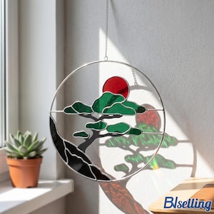 Könnte beinhalten: Runder Glas-Suncatcher mit einem stilisierten Baum und roter Sonne. Das Design umfasst grüne Blätter, einen schwarzen Berg und einen silbernen Metallrahmen. Aufgehängt an einer Kette.