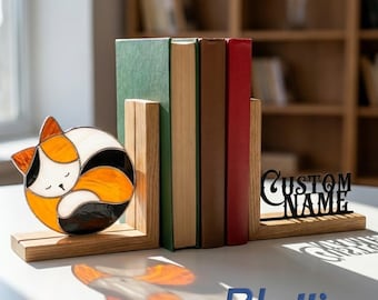 Puede incluir: Un juego de sujetalibros de madera con un gato de vidrieras en naranja, blanco y negro, y un segundo sujetalibros de madera con el texto CUSTOM NAME en letras negras, sosteniendo cuatro libros con lomos verde, beige, marrón y rojo.