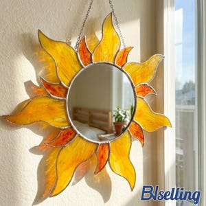 May include: A decorative sun-shaped mirror with a stained glass design. The mirror has a round center and is surrounded by yellow and orange glass flames. It hangs from a silver chain and is a unique wall decor piece.