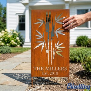A hand is holding a wooden sign with a bamboo design and the text "THE MILLERS Est. 2010" on it, standing in front of a white house.