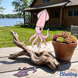 May include: A pale pink stained glass squid ornament mounted on a thin metal rod, which is inserted into a piece of weathered driftwood. The squid features a smooth, translucent finish with a single dark eye and multiple curved tentacles.