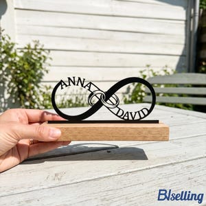 Puede incluir: Un recorte negro con forma de símbolo de infinito que integra los nombres ANNA y DAVID, junto con dos formas de anillos entrelazados. El recorte está montado sobre una base rectangular de madera clara, creando un letrero decorativo de mesa.