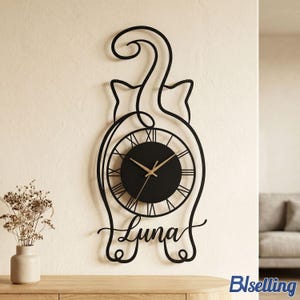May include: Black cat-shaped wall clock with a circular clock face featuring Roman numerals. The clock hands are gold. The name "Luna" is written in cursive below the clock face. The clock is mounted on a cream-colored wall.