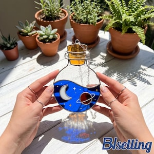 Puede incluir: Botella de poción de vidriera con un diseño celestial. La botella presenta un líquido azul con una luna creciente blanca, estrellas y un planeta. Tiene un corcho dorado y está suspendida por una cadena. El fondo incluye plantas en macetas.