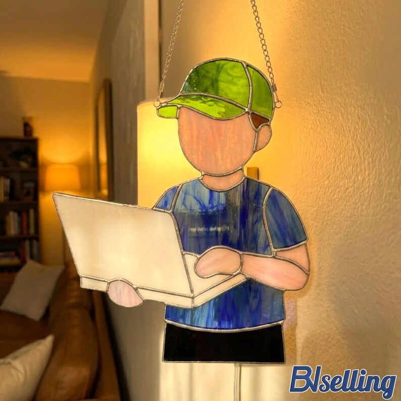 May include: Stained glass panel depicting a person wearing a green cap and blue shirt, holding a laptop. The panel is suspended by a chain. The figure's face and hands are pink, and the laptop is white.