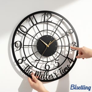 May include: A black, circular wall clock with gold hands and a black face. The clock features a unique open design with large, stylized numbers and the text "The Miller's Time" at the bottom. The clock is being held up by two hands.