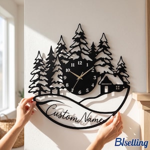 Könnte beinhalten: Eine schwarze Wanduhr mit einer lasergeschnittenen Silhouette eines Waldes mit Tannen und einer kleinen Hütte. Das runde Zifferblatt zeigt weiße Zahlen von 1 bis 12 mit goldenen Zeigern. Unter der Uhr steht auf einem geschwungenen Banner Custom Name.