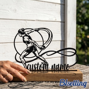 May include: Black metal sculpture of a discus thrower in action, mounted on a wooden base. The sculpture features a stylized athlete within a circular design, with the words "custom name" in cursive. The base is rectangular and made of light-coloured wood.