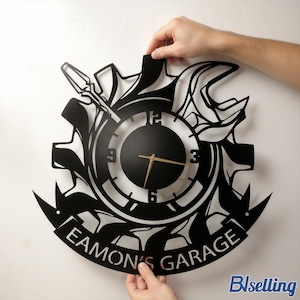 Peut inclure: Horloge murale en métal noir en forme d'engrenage, avec un cadran et des aiguilles dorées. L'horloge comprend une clé et un tournevis, avec le texte "Eamon's Garage" en bas. L'horloge est tenue par deux mains.
