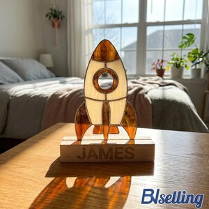 May include: A stained glass rocket ship ornament featuring amber and cream-colored glass panels with a circular window. The rocket is mounted on a rectangular wooden base with the name JAMES engraved in capital letters on the front.
