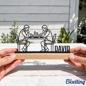 May include: A black metal silhouette of two people sitting at a table playing chess, mounted on a rectangular light wood base. The design features the name DAVID in bold black capital letters integrated into the right side of the structure.