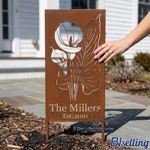 A hand is holding a wooden sign with a metal cutout of a flower and the text "The Millers Est. 2010" on it, standing on a bed of mulch.
