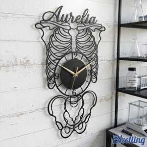 May include: Black metal wall clock with a skeleton design, featuring the name "Aurelia" at the top. The clock has a round black face with gold hands. The design includes a rib cage, spine, and pelvis. The clock is mounted on a white wooden wall.