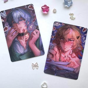 May include: Two rectangular art prints featuring anime-style illustrations. The left print shows a character with blue hair, a collar, and a green apron. The right print depicts a character with brown hair, horns, and red markings on their chest.