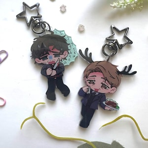 May include: Two acrylic keychains with star-shaped clasps. One features a brown-haired character holding a knife with a clock-like background. The other shows a light-brown-haired character with antlers holding a plate of food. Both have chibi-style art.