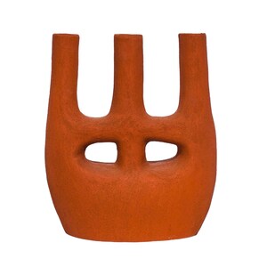 May include: A terracotta-colored ceramic vase with a textured, matte finish. The vessel features a wide, rounded base that transitions into three distinct, upright cylindrical openings at the top, separated by two horizontal oval cutouts.