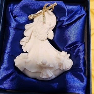 Op de afbeelding: Een crèmekleurig engelenbeeldje met een gouden lint, gepresenteerd in een blauw satijn gevoerde doos. De engel is gebeeldhouwd met vloeiende gewaden en heeft delicate details. Het beeldje is een religieus of decoratief item.