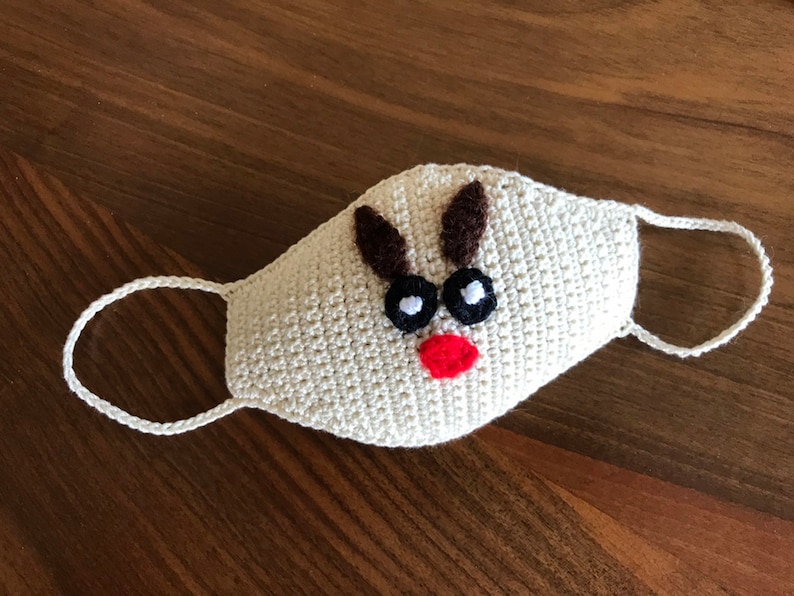 Puede incluir: M&aacute;scara facial de crochet blanca con un dise&ntilde;o de reno. La m&aacute;scara tiene cuernos marrones, ojos negros y una nariz roja.