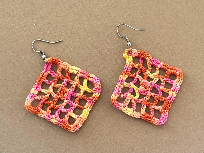 May include: A pair of crocheted earrings with a square shape. The earrings are made with a multi-colored yarn in shades of orange, pink, and yellow.