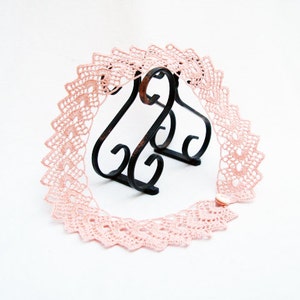 May include: A delicate pink crocheted lace trim with a scalloped edge. The lace is displayed on a black metal stand.