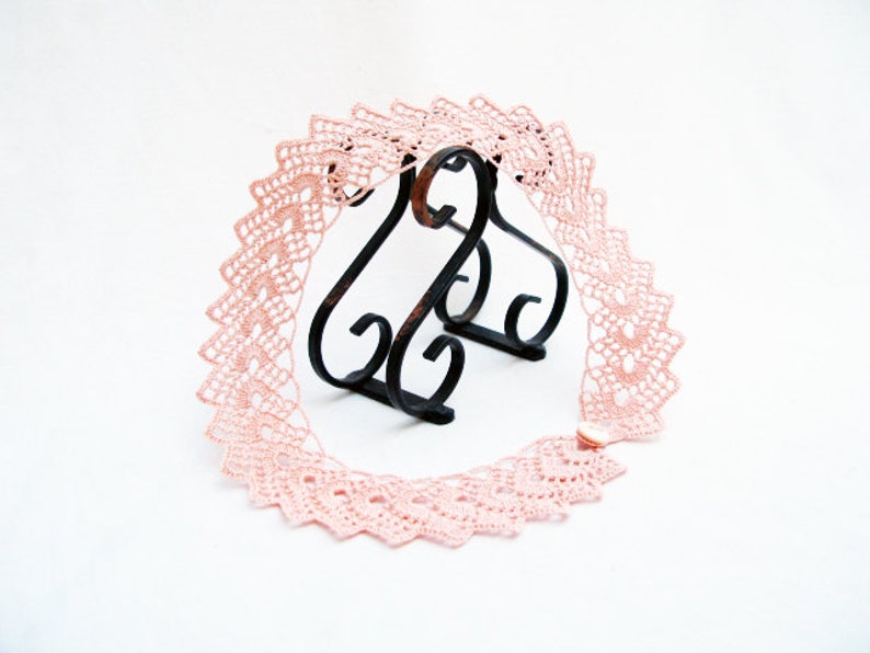 May include: A delicate pink crocheted lace trim with a scalloped edge. The lace is displayed on a black metal stand.